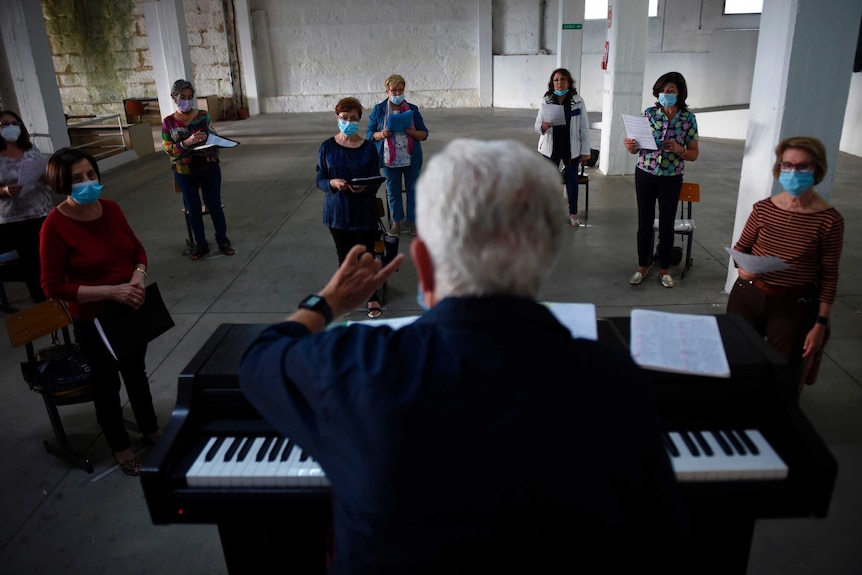 A choir director stands at a piano with a group of choristers spread out in front of him