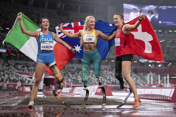Three women with lower limb different stand on a podium waving their national flags.