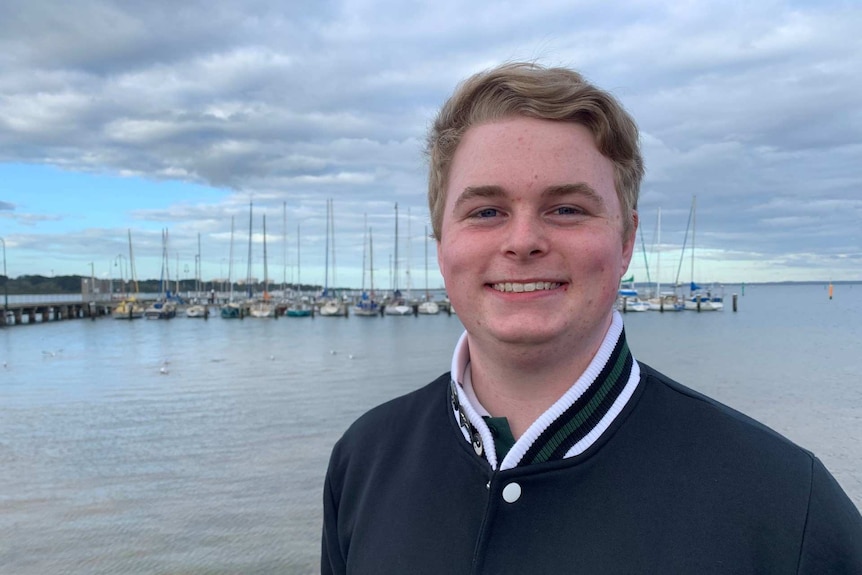 Ryan Hicklin stands at the beach with sailing boats in the background.