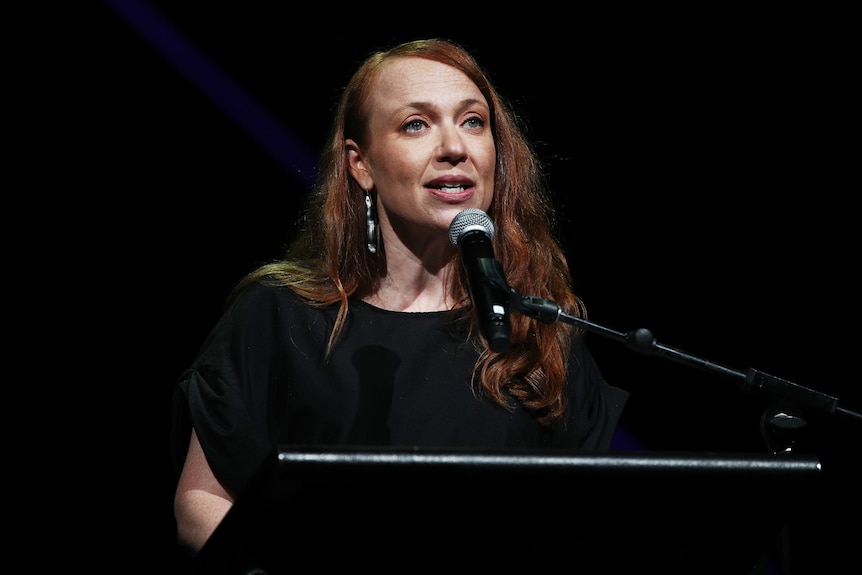A middle-aged woman with auburn hair wearing a black shirt stands at a podium, speaking into a microphone