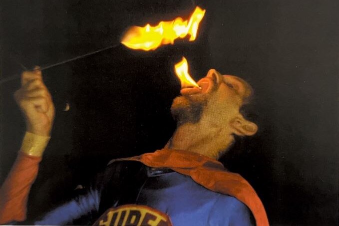 A man with fire on his tongue, holding a stick with a fire burning on one end. 