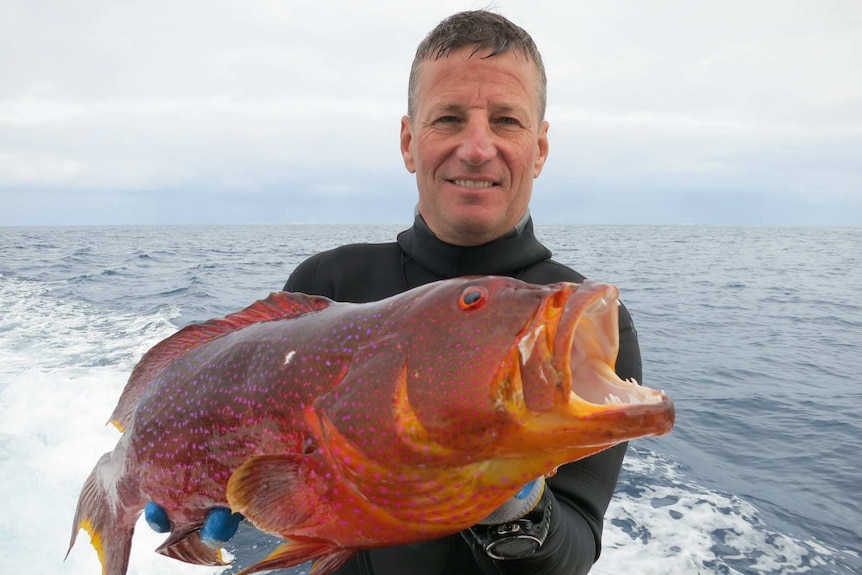 A white man wearing a wetsuit holds up a large orange fish.