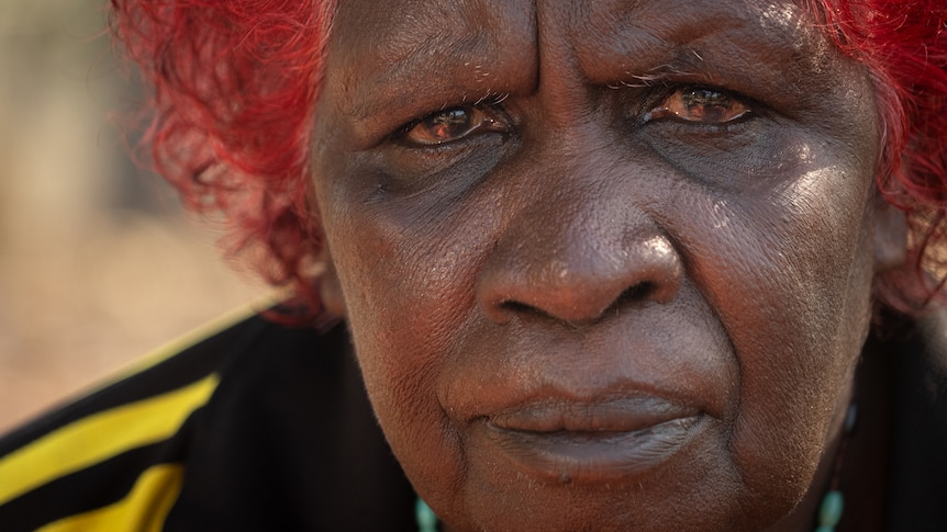 A middle-aged Indigenous woman with dyed red hair looks at the camera with a neutral expression.