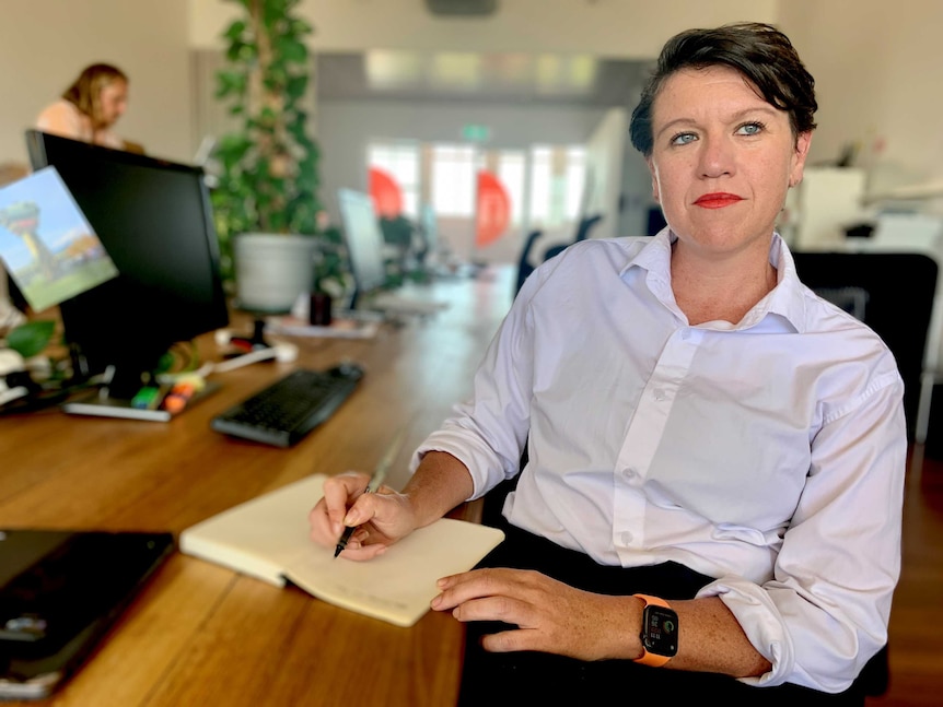 Brynn O'Brien sits at her desk with a pen in hand and a computer and colleague in the background.