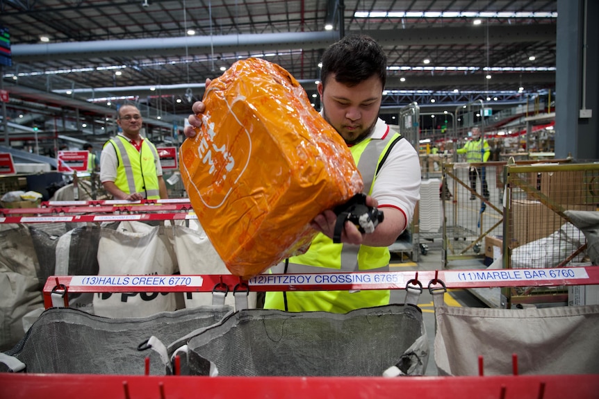 A young man holding a a large orange parcel over a sorting bin in an Australia Post warehouse, with and older man behind right.