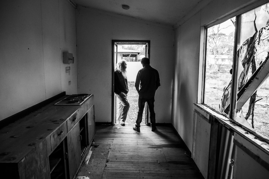 A black and white photo showing an old kitchen. Two men stand talking at the far end.