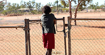 An Aboriginal boy stands at a fence with his back turned to the camera.