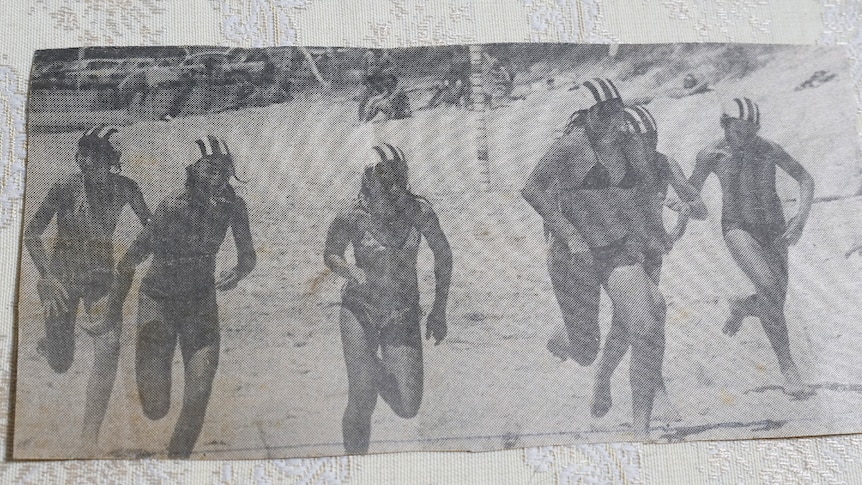 A newspaper clipping of teenage girls running on the sand, wearing striped caps showing which surf club they belong to.