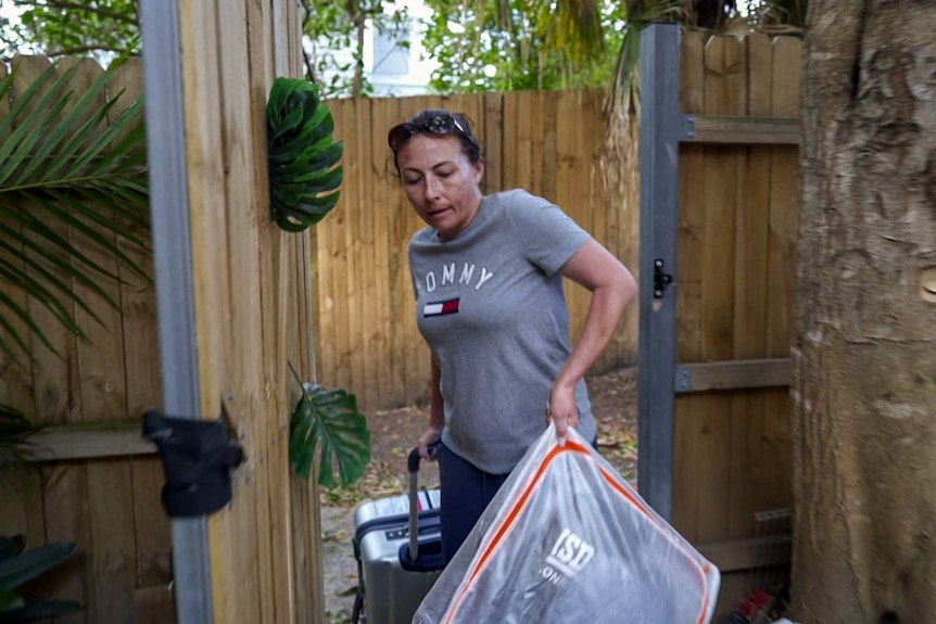 Woman wearing grey top carries two bags through an outside wooden gate