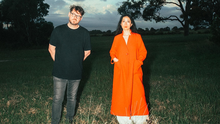 Mindy Meng Wang 王萌 and Tim Shiel stand in a field at dusk. She wears a red coat, he wears a black t-shirt
