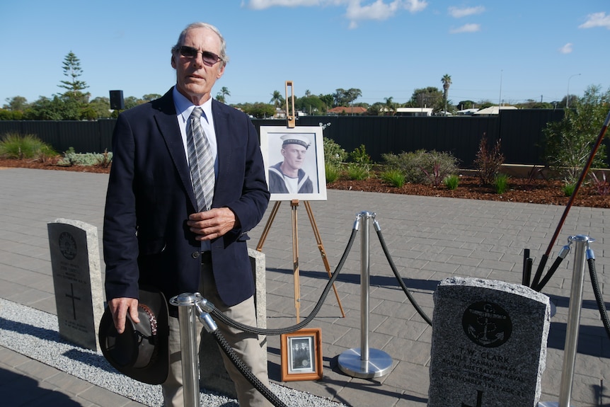 Man in a blazer stands beside a headstone with a black and white photograph on an easel behind it.