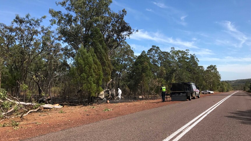 Police and forensic officers on the side of the road where a burnt out car lies in the bush.