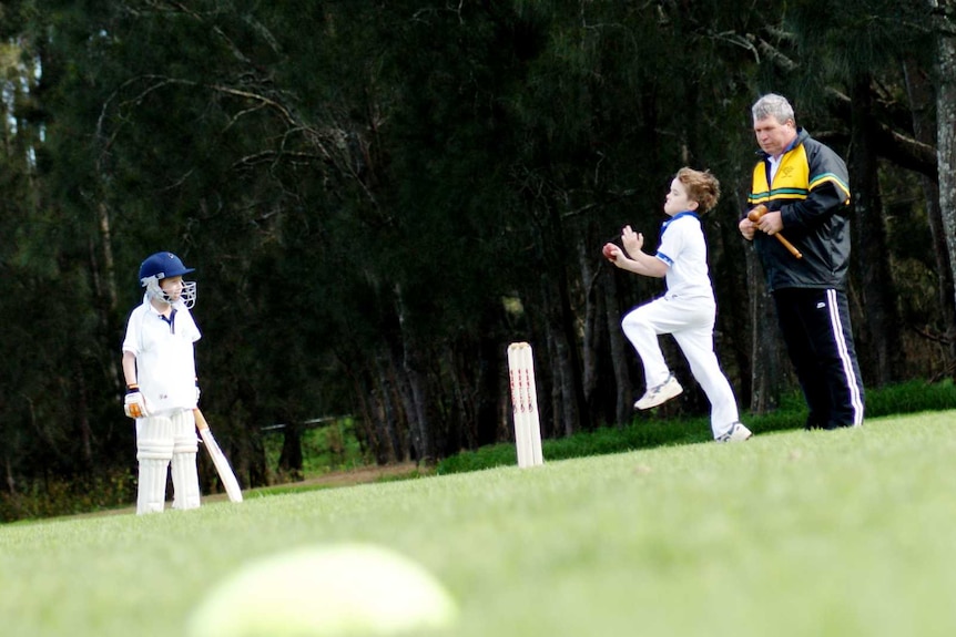 A young boy runs in to bowl during a junior cricket game as a batter and umpire in tracksuit look on