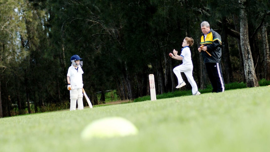 A young boy runs in to bowl during a junior cricket game as a batter and umpire in tracksuit look on