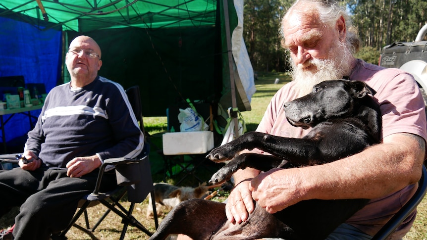 Two older men sitting in camp chairs, one with a medium-sized black dog in his lap