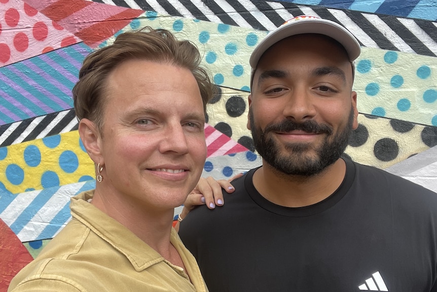 Shane, left, smiles as he puts a hand on Alex's shoulder, right, while they both stand in front of a mural on a sunny day.