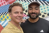 Shane, left, smiles as he puts a hand on Alex's shoulder, right, while they both stand in front of a mural on a sunny day.