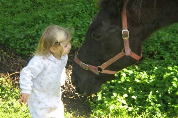 Kyanne standing on grass, blonde hair half tied up in a one piece, leaning into a horse face.