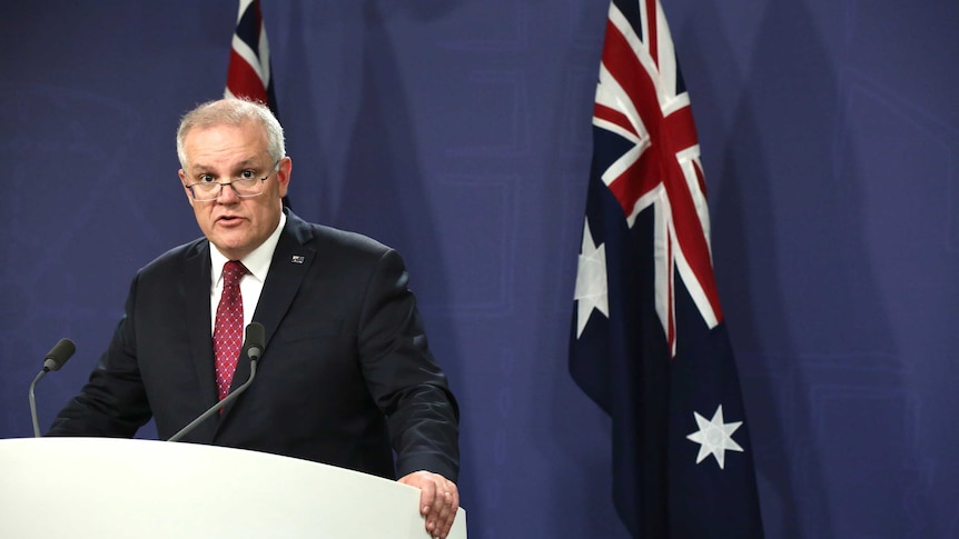 A man with grey hair and glasses speaking a lecturn in front of two australian flags