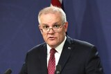 A man with grey hair and glasses speaking a lecturn in front of two australian flags