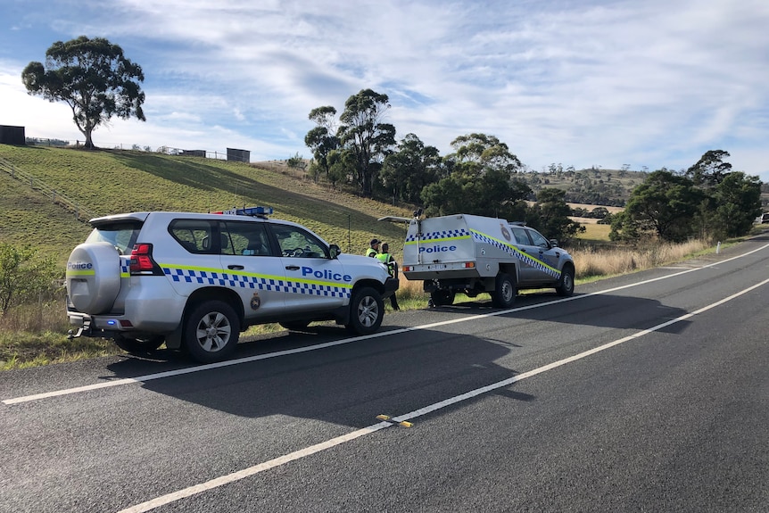 Police stand near two police cars on the side of a road.