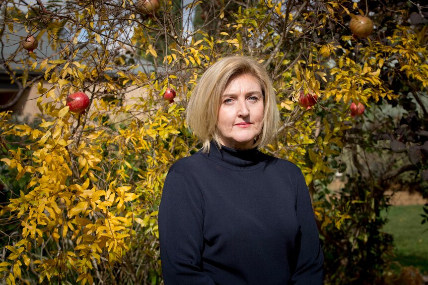 A middle aged white woman with short, fair hair stands in front of a deciduous tree