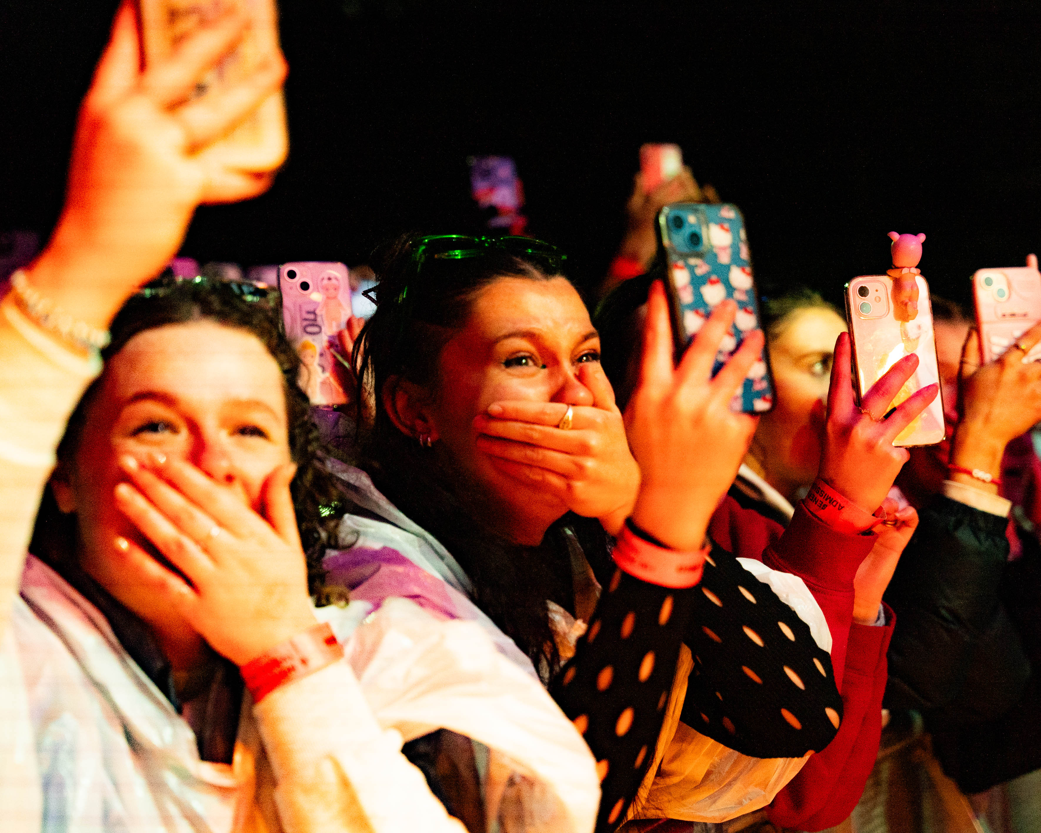 A crowd of people hold their phones up recording a live gig. Two people have their hands over their mouths in happiness