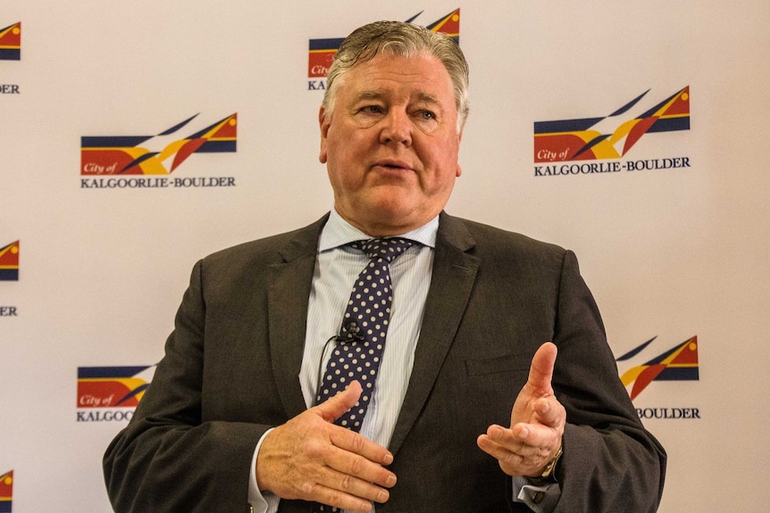 A man in a suit talking in front of a billboard with the name of his employer.  