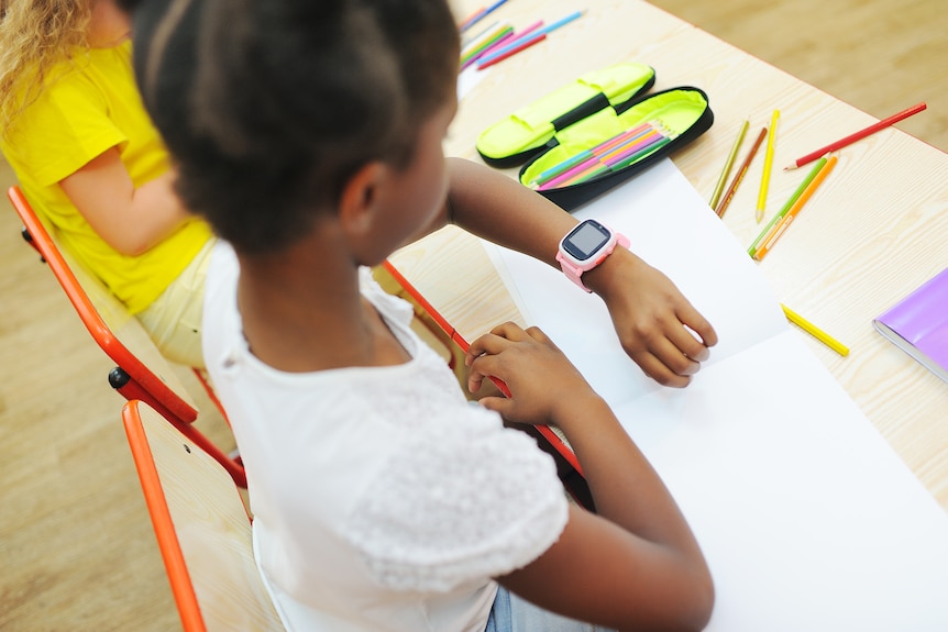 a young girl with black hair and white top sits at a school desk with paper and pencils as she looks at a pink smartwatch