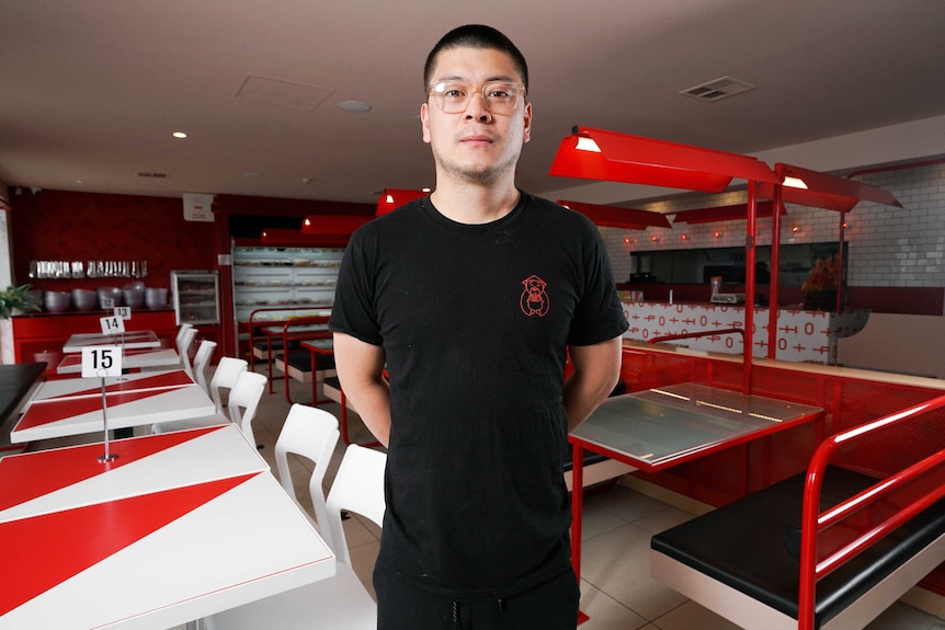 A man dressed in black wearing glasses stands in the foreground of an empty restaurant.