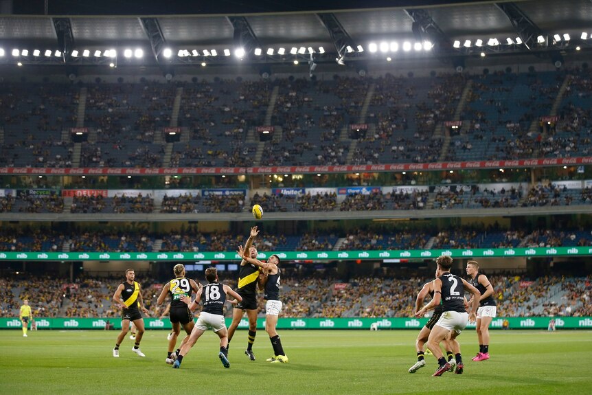 Richmond and Carlton players jostle for the ball in front of a crowd during a night match at the MCG.