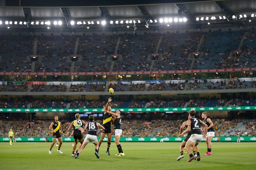 Richmond and Carlton players jostle for the ball in front of a crowd during a night match at the MCG.
