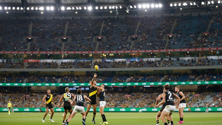 Richmond and Carlton players jostle for the ball in front of a crowd during a night match at the MCG.