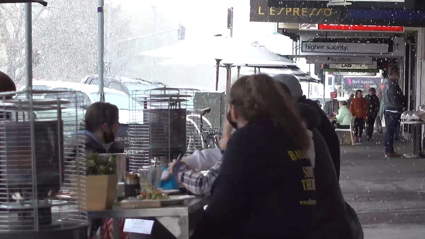 Main street of Ballarat with snow flakes falling down, people sitting on outdoor cafe tables with heaters, others walking.