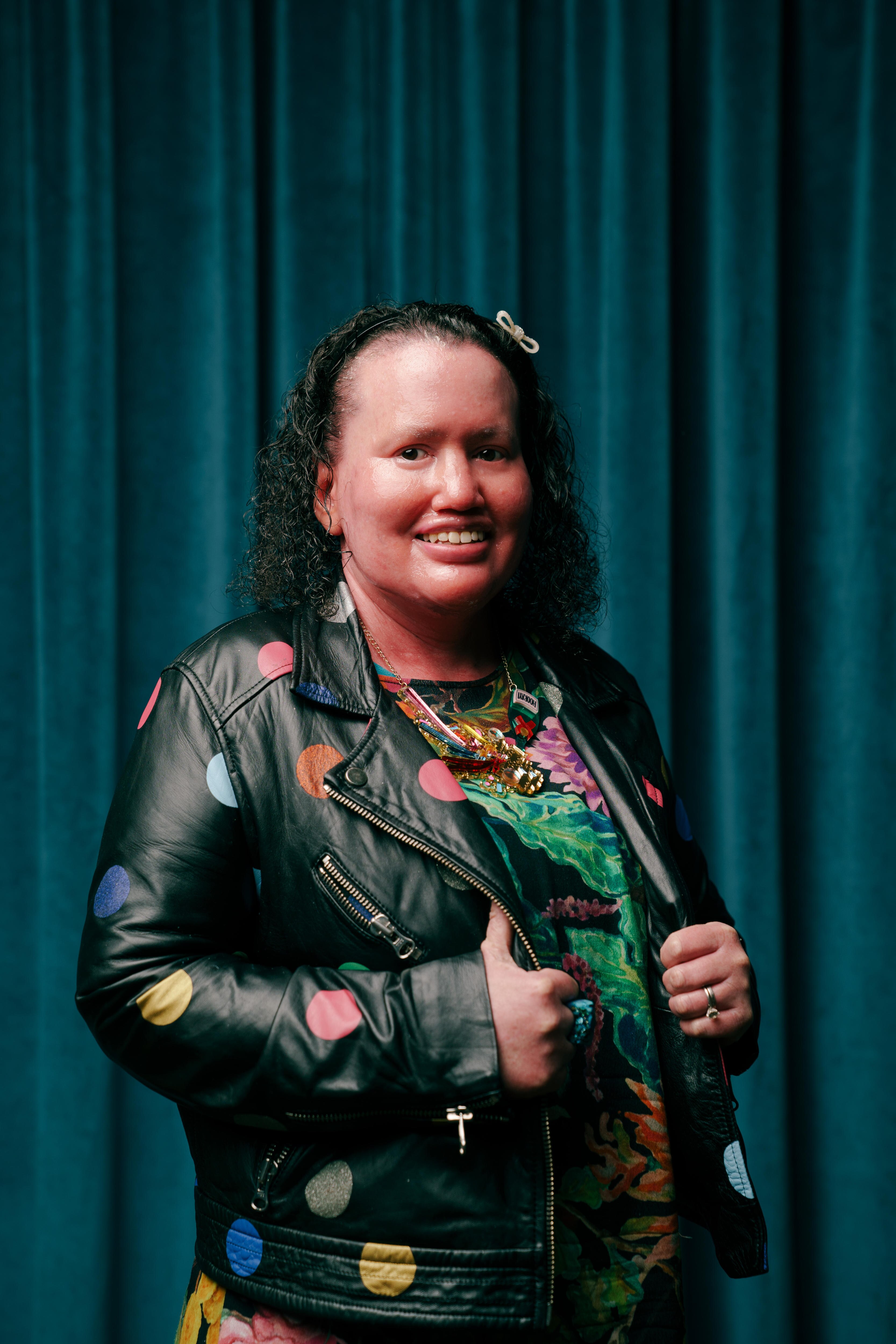 A woman with curly hair, wearing a bright dress and polka-dot leather jacket stands smiling in front of a blue velvet curtain.