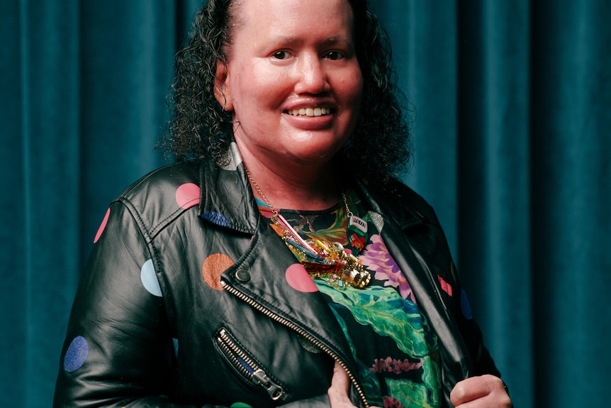 A woman with curly hair, wearing a bright dress and polka-dot leather jacket stands smiling in front of a blue velvet curtain.