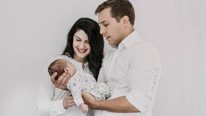 A man and a woman smile as they hold their newborn child. All three are dressed in white.