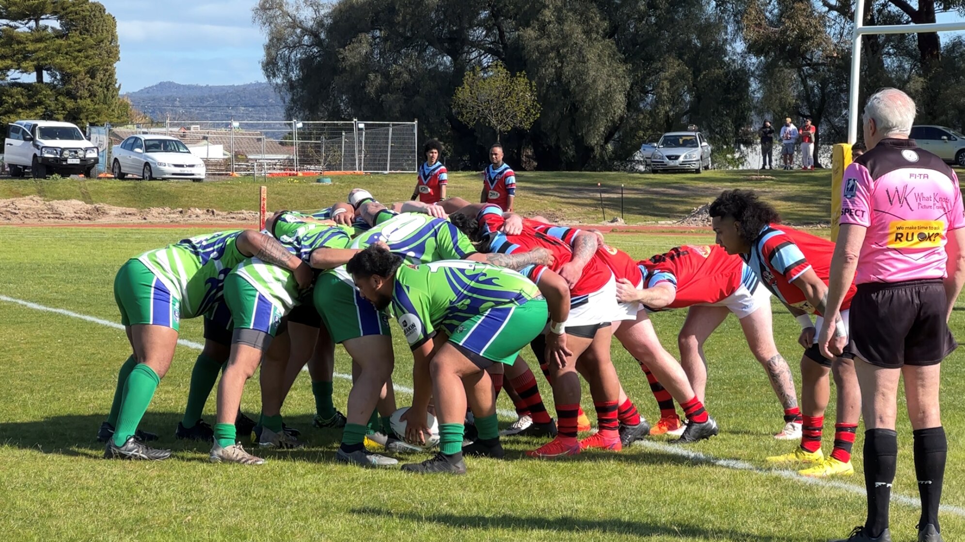 A rugby scrum with a team in green shirts, shorts and socks, and another in red and blue shirt white shorts. ref in pink 