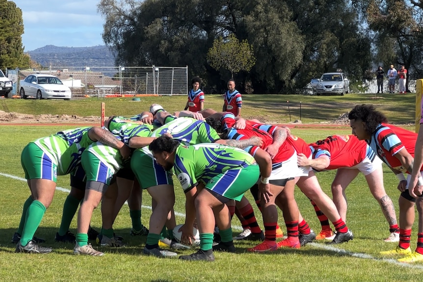 A rugby scrum with a team in green shirts, shorts and socks, and another in red and blue shirt white shorts. ref in pink 