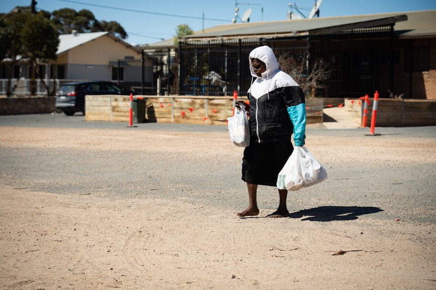 An Aboriginal woman carrying shopping bags walks across a dusty road