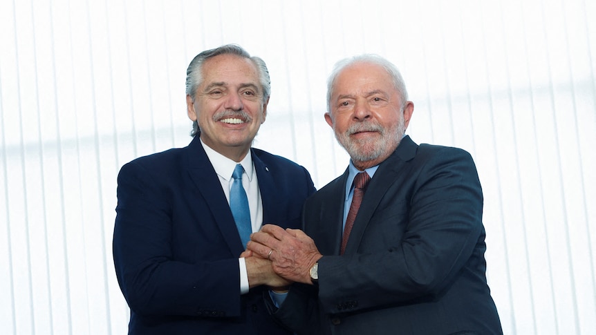 Two older hispanic men in dark suits clasp hands as they stand in front of well-lit white shutters.