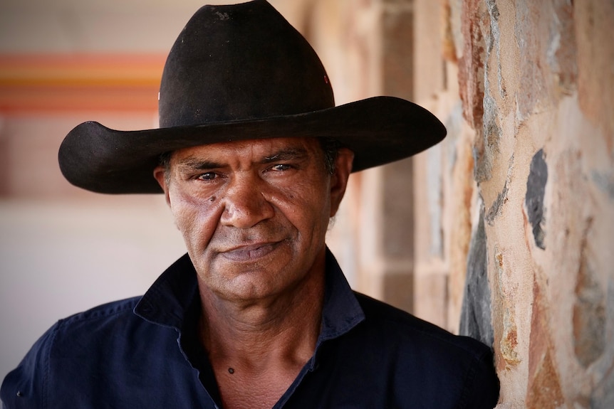 Portrait of an Aboriginal man wearing black wide-brimmed hat standing against rock wall of a hotel