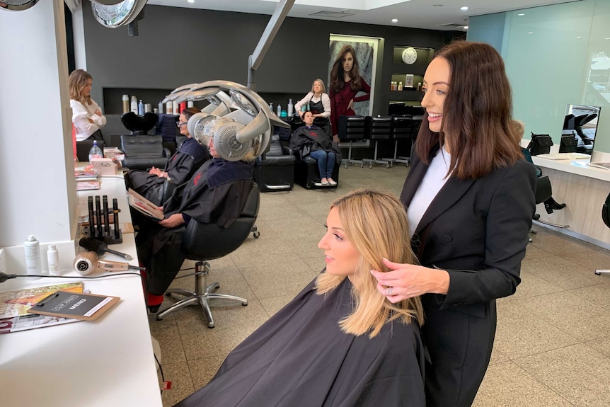 A women in a smock at a hairdressing salon with another woman behind her touching her hair