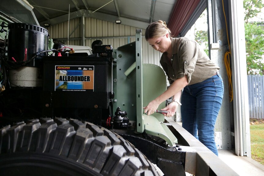 The side view of a vintage four-wheel drive with exposed engine, on the left side a woman is working on the vehicle.