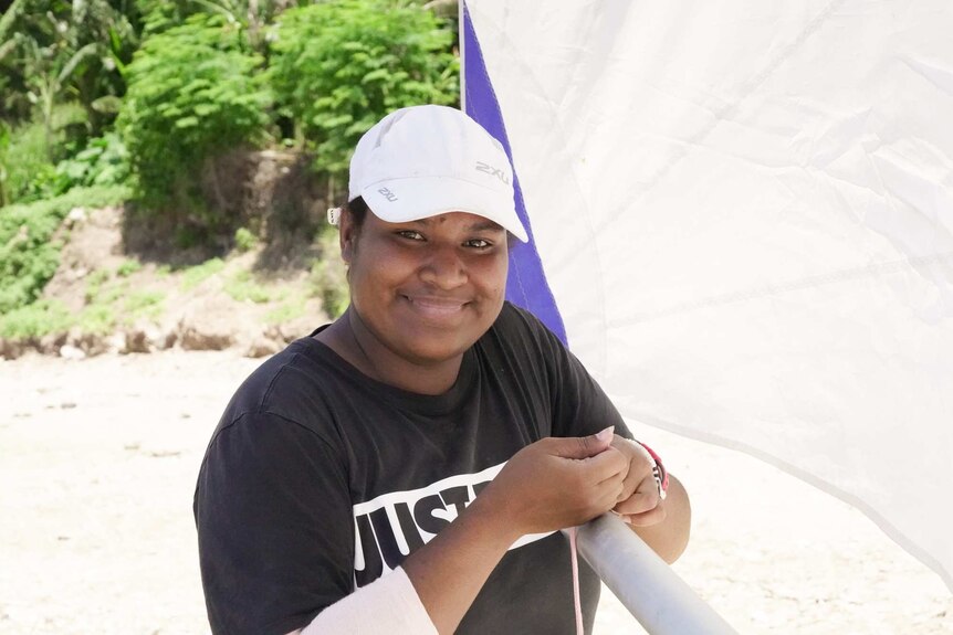 A Black woman wearing a white cap and a black shirt smiles as she leans on her sail.