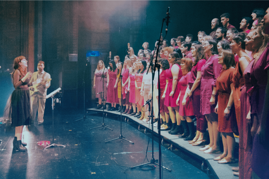 a woman stands on a stage directing a large choir of people wearing bright colours.