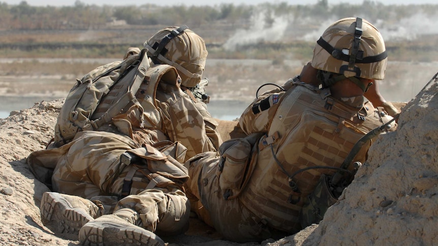 Two armed soldiers in full kit look over an arid valley, one aims a sniper rifle while the other scans the horizon