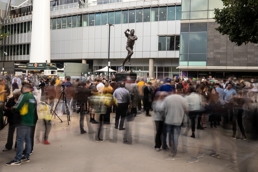 Crowds gather around a statue of Shane Warne.