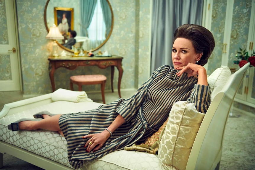 Naomi Watts as Babe Paley poses for a photo on a sofa wearing a brunette beehive hairstyle and a striped dress