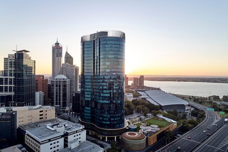 Woodside's Perth office tower, a glass-fronted building towering over the sea and other buildings, sunrise in the horizon.